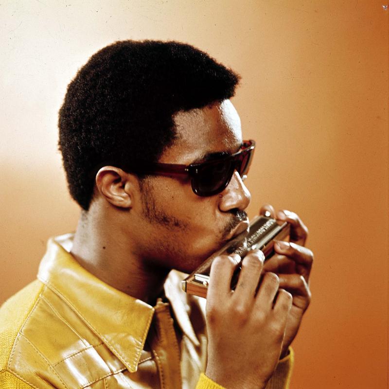 A young Stevie Wonder, pictured during his Motown days, plays the harmonica in sunglasses against an orange backdrop